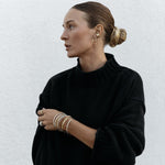 A woman wearing multiple stacked Flirt Bangles in gold and silver, a white gold Etched Paris Ring, an Open Diamond Ring and a C’est Moi Link Ring, two Ready to Mingle Ear Cuffs, Sleek & Chic Hoops and an Oval Hoop, all earrings in yellow gold, styed with her hair in a bun and a black knit.