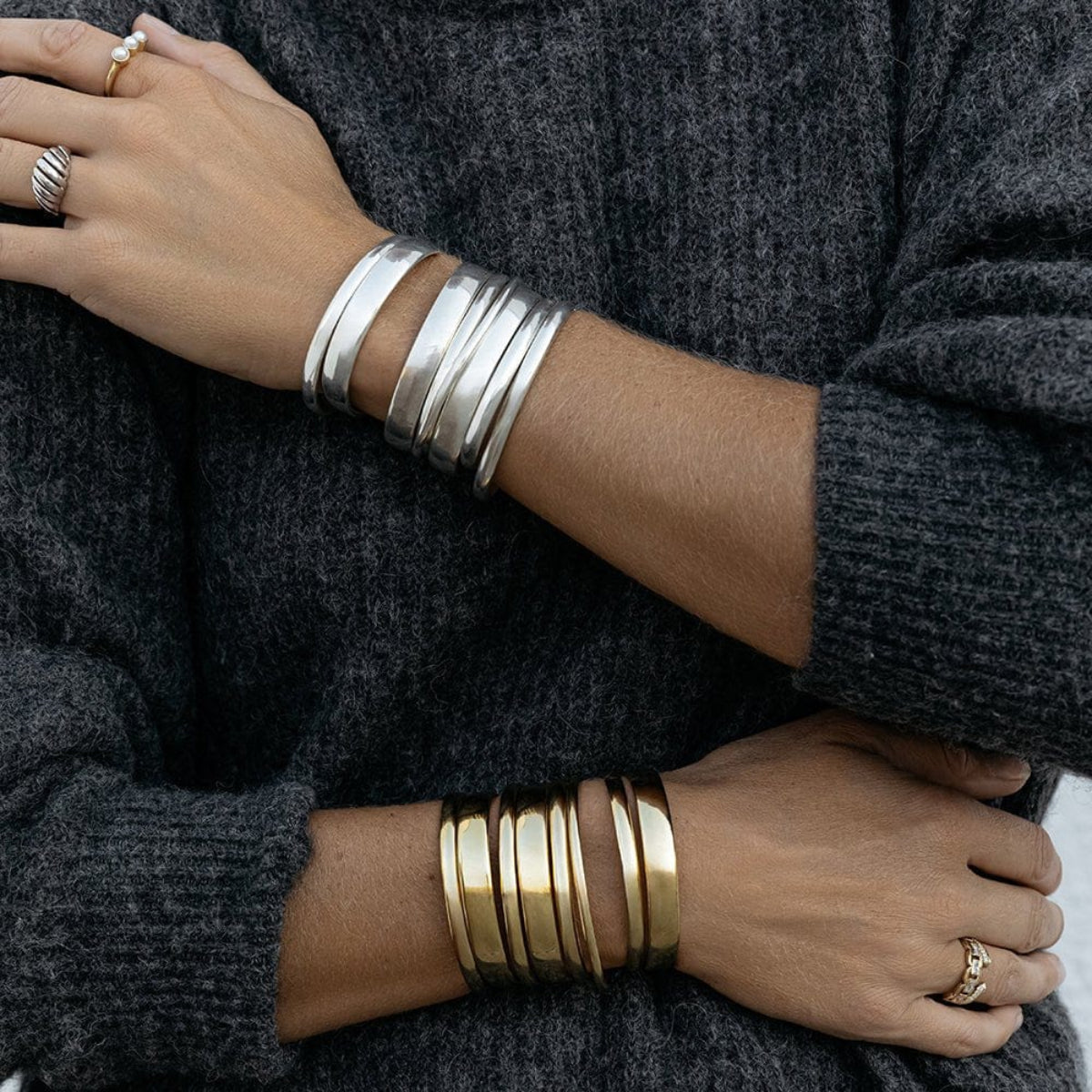 A close-up of arms wearing stacked Rendezvous Cuffs, Plus One Cuffs and All In Cuffs, one arm in silver and the other in gold, a Perla Pinky Ring, an Etched Paris Ring, a C'est Moi Link Ring and an Open Diamond Ring, styled in a dark grey knit.