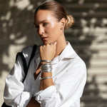 A woman wearing stacked sterling silver All In Cuffs and Plus One Cuffs, an Etched Paris Ring, a Ready to Mingle Ear Cuff and Sleek & Chic Hoops, with her hair in a bun and styled in a white shirt.