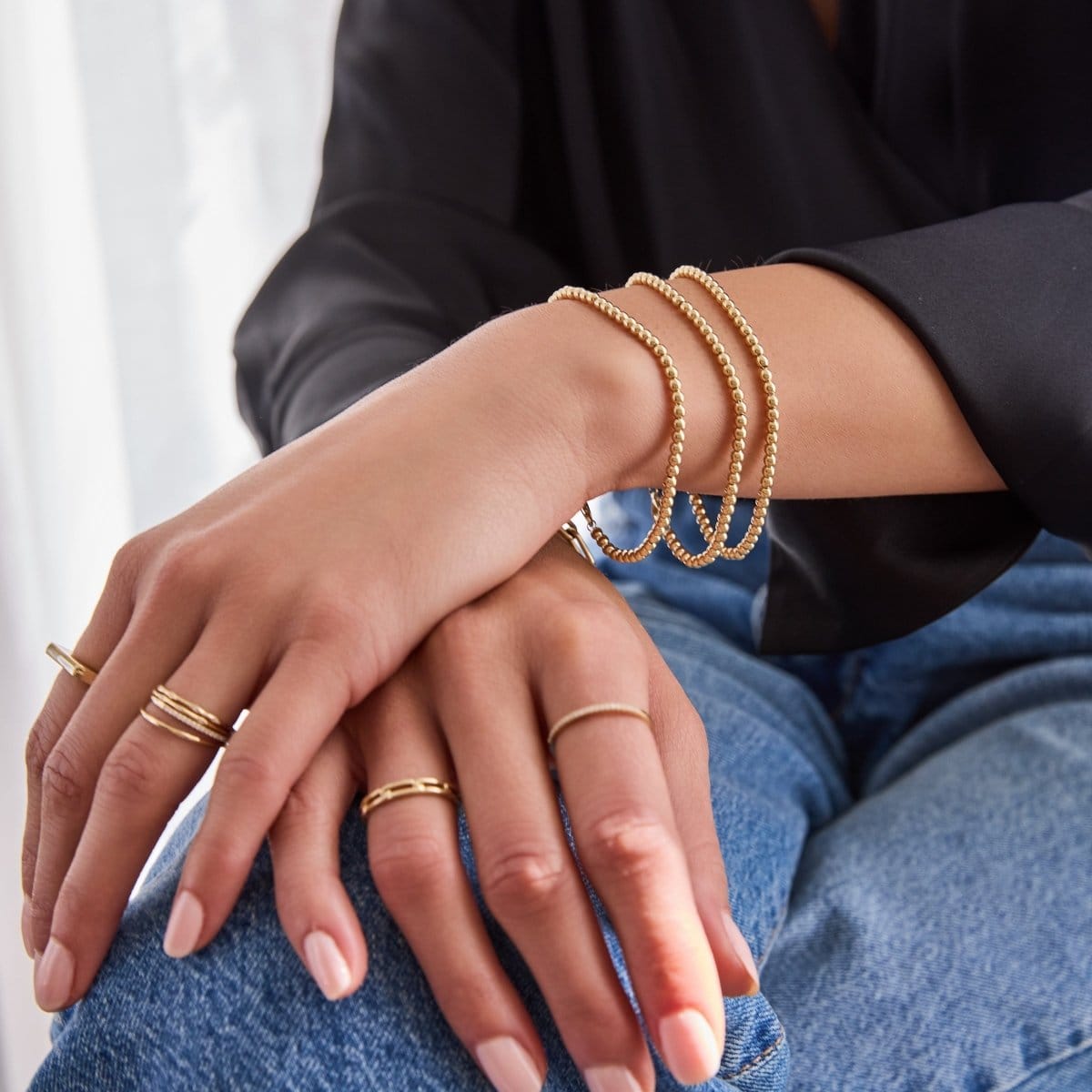 A close-up of a woman's hands wearing three stacked Bubble Bracelets in small, and various yellow gold rings, including a Link Ring, Delicate Bands, Pavé Diamond Rings, a 4 Diamond Ring and a Geo Stacking Ring, styled in blue jeans and a black silk top.