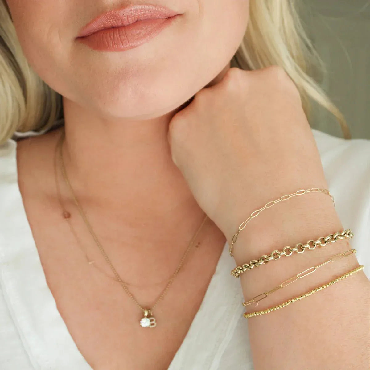A close-up of a woman’s neck and wrist wearing an Element Short Link Bracelet, an Element Long Link Bracelet, a Rolo Bracelet and a Diamond Cut Bracelet, a Custom Gold Necklace with 2 letters (an L and a birthstone charm), and a Classic Bead Chain with a Diamond Letter Jumble featuring the letter B, all in yellow gold, styled with a white top.