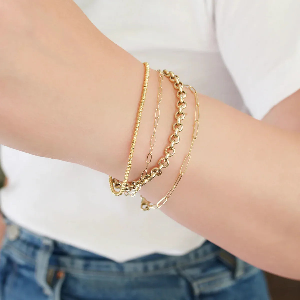 A close-up of a woman’s arm wearing yellow gold bracelets: a Diamond Cut Bracelet, an Element Long Link Bracelet, an Element Short Link Bracelet and a Rolo Bracelet, with a white top and blue jeans.