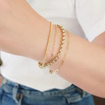 A close-up of a woman’s arm wearing yellow gold bracelets: a Diamond Cut Bracelet, an Element Long Link Bracelet, an Element Short Link Bracelet and a Rolo Bracelet, with a white top and blue jeans.