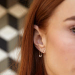 Close-up of a woman's ear wearing a Pavé Safety Pin Earring, a 14K Gold Number Stud in 3 and a 14k Gold Pavé Letter Stud, with her orange hair visible against a blurred patterned background.