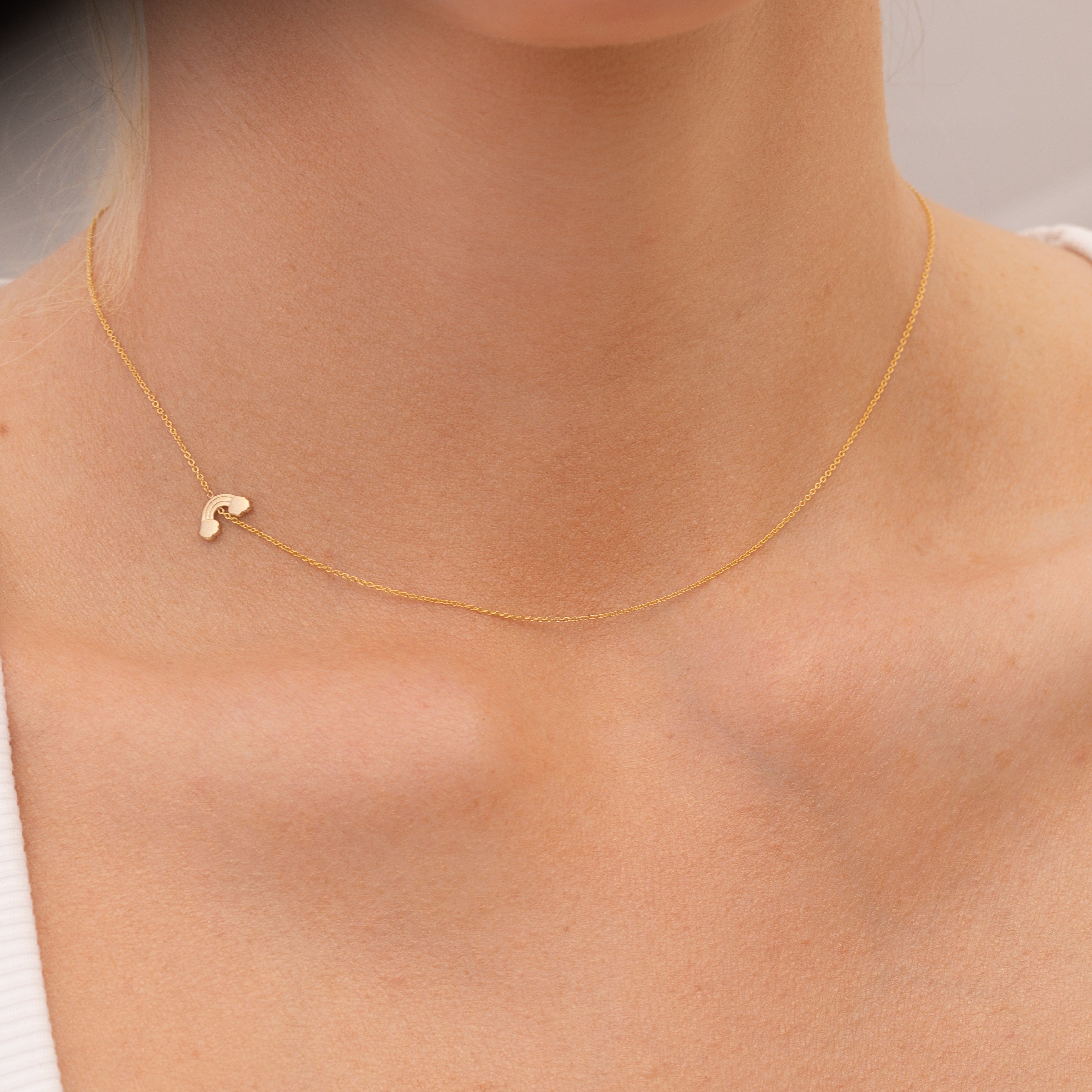 Gold necklace with a small rainbow charm on a person's neck against a neutral background