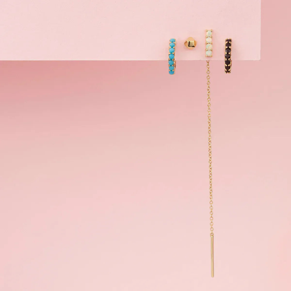 An Alli Webb Turquoise Huggie, Faceted Comfort Stud, Opal Threader and Onyx Huggie on a light pink background. 