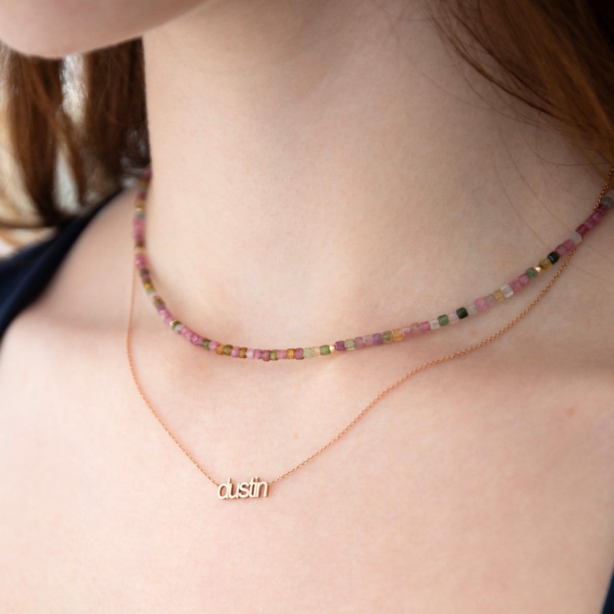 Close-up of a neck wearing an Endless Summer Necklace - Coral Reef and a Petite Nameplate Necklace with 'dustin' in rose gold. 