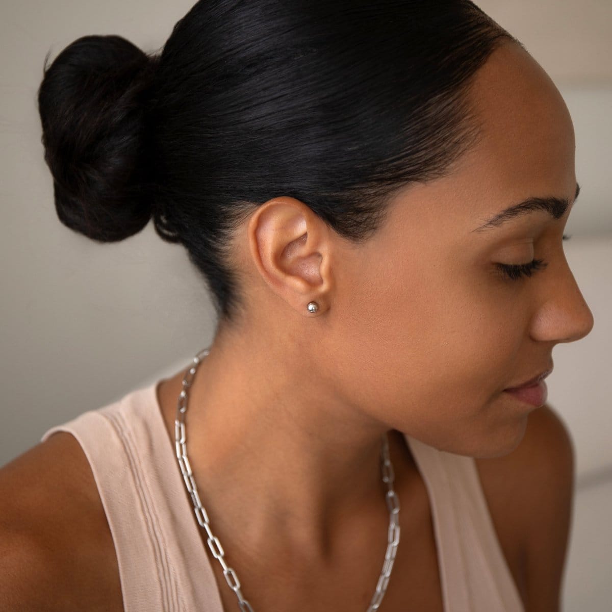A woman wearing a white gold Preppy Post and a Bold Link Chain with a nude tank top and her black hair in a bun, against a light grey background.