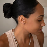 A woman wearing a white gold Preppy Post and a Bold Link Chain with a nude tank top and her black hair in a bun, against a light grey background.