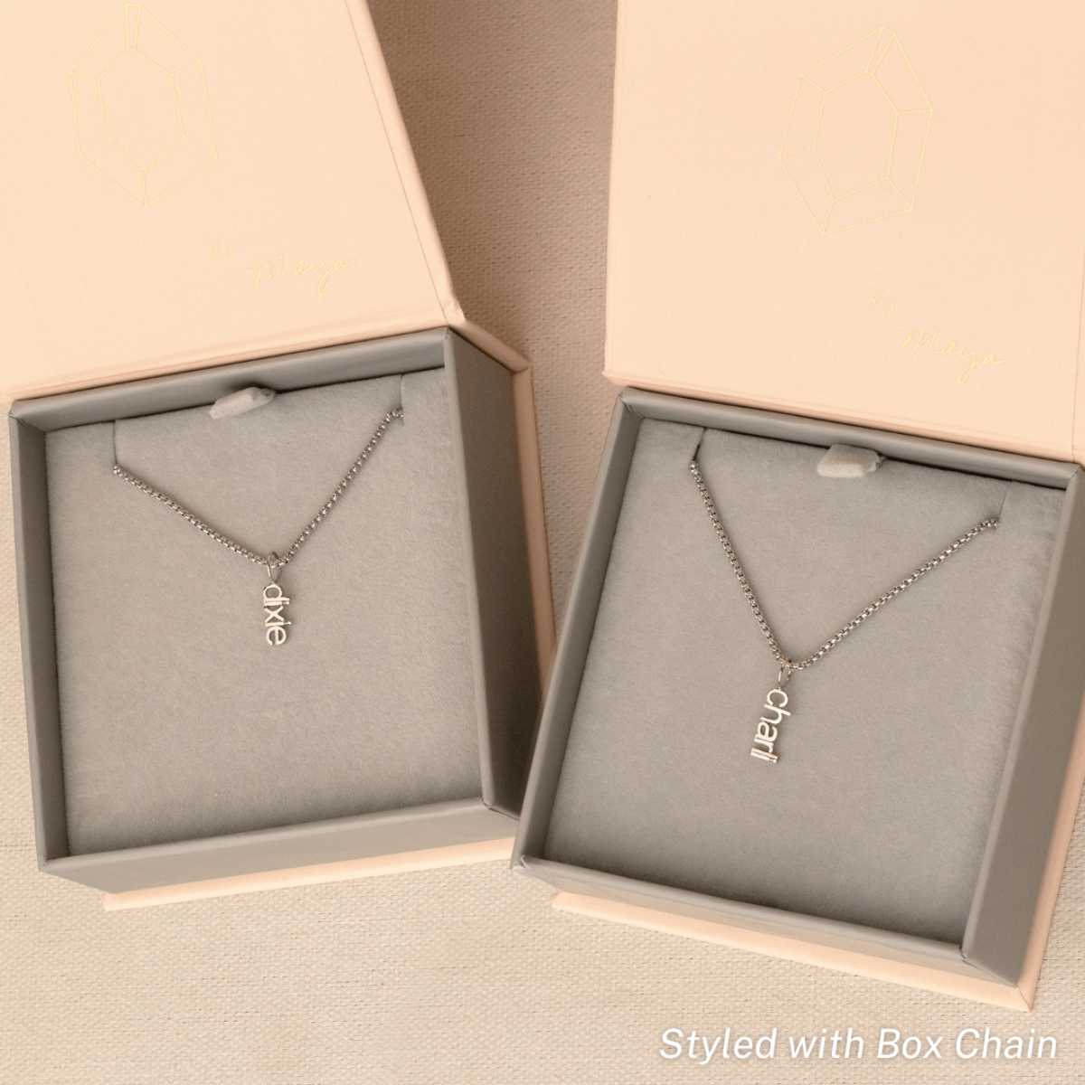 Two white gold Box Chain necklaces each with a Petite Nameplate Pendant - one reading ‘dixie’ and the other ‘charli’, placed in separate light grey and light pink jewellery boxes on a beige background.