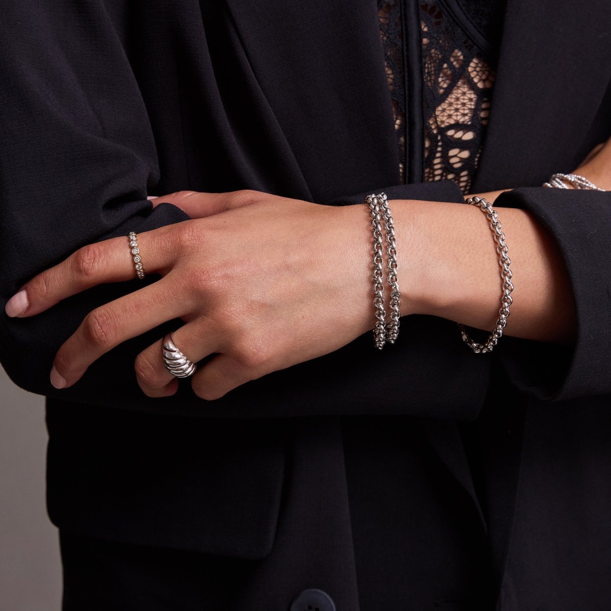 A close-up of a woman’s hand wearing three Rolo Bracelets, an Evermore Ring and an Etched Paris Ring, all in white gold, with a navy suit and lace top.