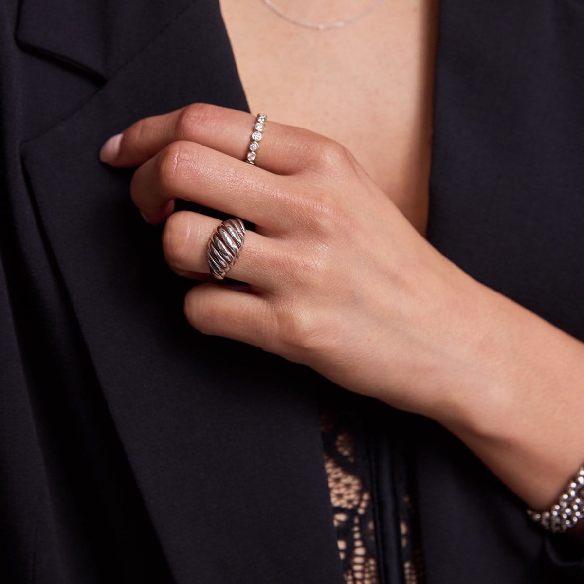 A woman's hand wearing a white gold Etched Paris Ring, The Evermore Ring and Rolo Bracelets, styled in black blazer and lace top. 