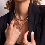 Close-up of a woman's neck wearing white gold jewellery - a Rolo Necklace, a Sequence Chain, a 14k Gold Asymmetrical Letter Necklace - T, a Sharktooth Necklace, a Gold Lariat Necklace, a Heartfelt Ring, an Etched Paris Ring, and two Evermore Rings, with a black lace top and blazer against a light grey background.