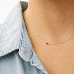 Necklace with a small heart pendant and red gemstone worn by a person in a denim shirt.