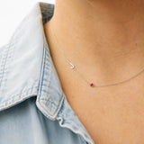 Necklace with a small heart pendant and red gemstone worn by a person in a denim shirt.