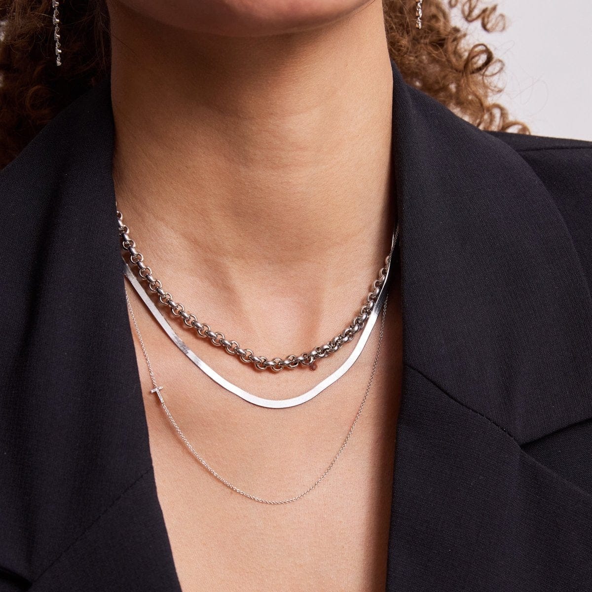 Close-up of a woman's décolletage wearing a Rolo Necklace, a Sequence Chain and a 14k Gold Asymmetrical Charm Necklace - Pavé Cross, all in white gold, with a black blazer and her brown curly hair partially visible.