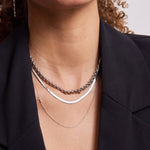 Close-up of a woman's décolletage wearing a Rolo Necklace, a Sequence Chain and a 14k Gold Asymmetrical Charm Necklace - Pavé Cross, all in white gold, with a black blazer and her brown curly hair partially visible.