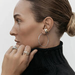 A woman wearing two Ready to Mingle Ear Cuffs, one in white gold and one in yellow gold, a Sleek & Chic Hoop - Medium, Etched Paris Ring, C'est Moi Link Ring and an Open Diamond Ring, with a black high-neck knit and her blonde hair in a bun against a white background. 