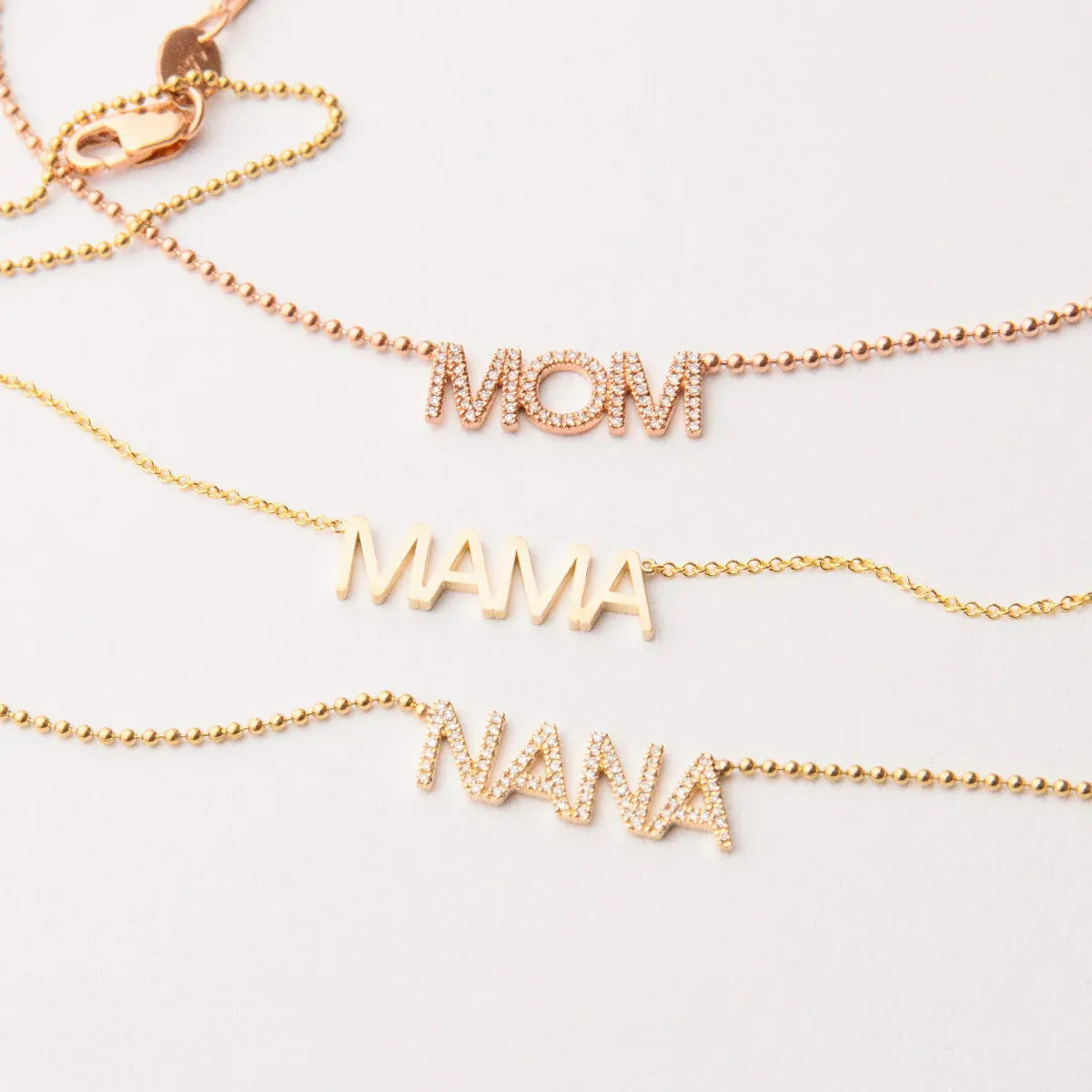 A Pavé MOM Necklace in rose gold, a MAMA Necklace in yellow gold and a  Pavé MOM Necklace in yellow gold, on an off-white background. 