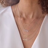 Close-up of a woman's décolletage wearing a MUM Necklace in rose gold, a Pavé MAMA Necklace in white gold, a MOM Necklace in yellow gold and a MOMMY Necklace in mixed metal, with a white top and brown curly hair. 