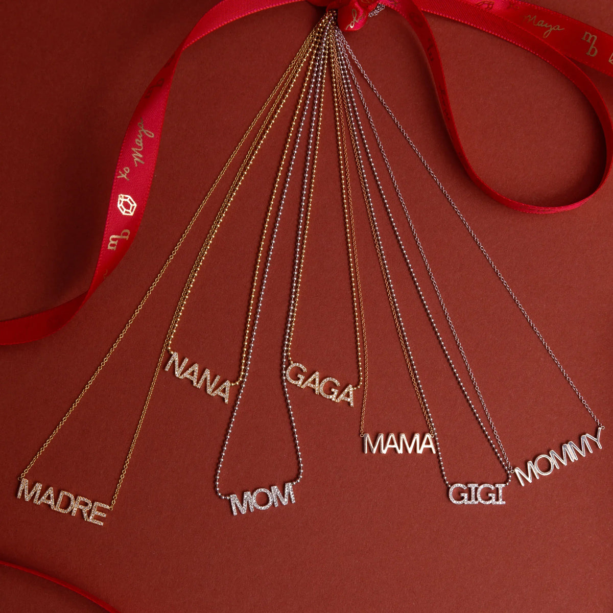 Pavé MADRE, Pavé NANA, Pavé GAGA, Pavé GIGI, MAMA, and MOMMY Necklaces tied to a red ribbon against a red background.