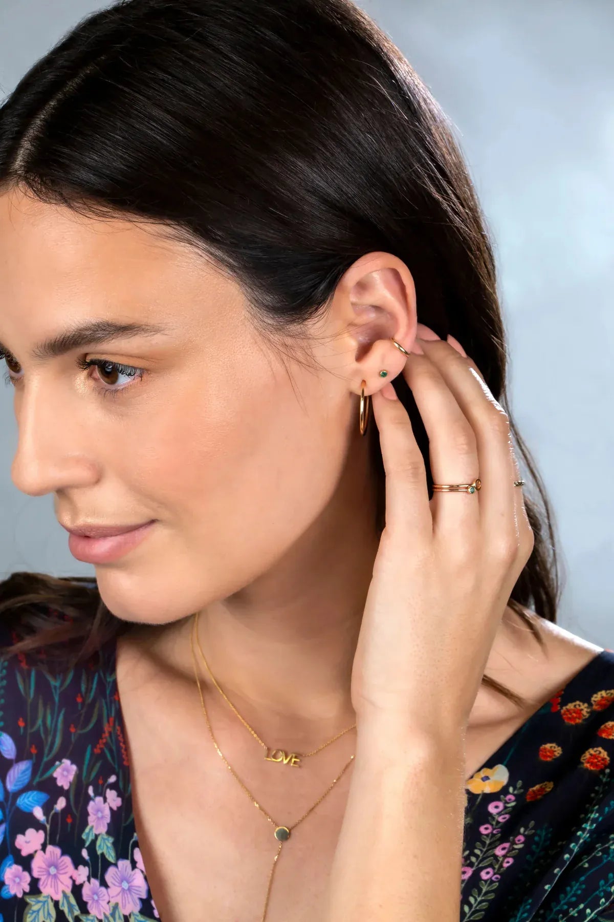 A Birthstone Stud, Rounded Ear Cuff, yellow gold hoop earring, two Birthstone Rings and LOVE Necklace worn by a brunette woman with a floral navy top against a light blue background. 