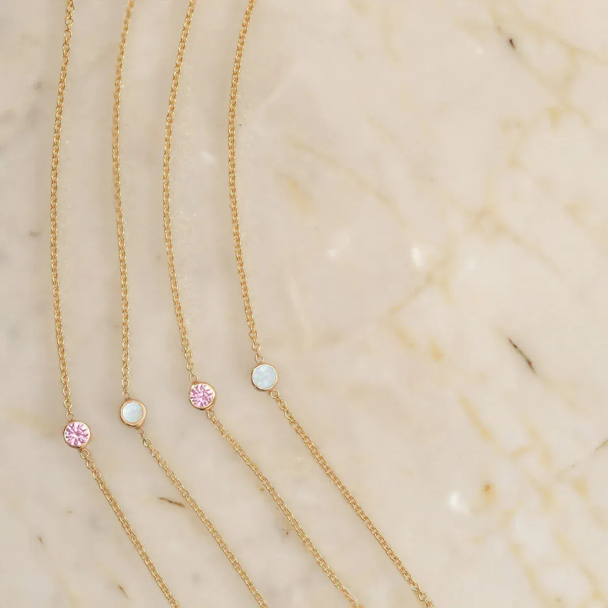 Four yellow gold 14K Gold Asymmetrical Birthstone Necklaces (two Opal and two Pink Tourmaline) on a light beige marble background. 