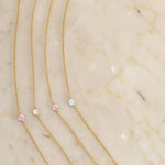 Four yellow gold 14K Gold Asymmetrical Birthstone Necklaces (two Opal and two Pink Tourmaline) on a light beige marble background. 