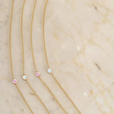 Four yellow gold 14K Gold Asymmetrical Birthstone Necklaces (two Opal and two Pink Tourmaline) on a light beige marble background. 
