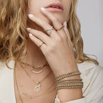 A woman with her hands wearing a Criss-Cross Diamond Daze Ring, Braided Ring, Birthstone Ring, Pave Geo Ring - Black Diamond and several Bubble Bracelets, along with a Bold Link Chain, Diamond Arc Layering Necklace, an Element Long Link Chain with a Diamond Pendant and Zodiac Pendant – Aries, two Custom Gold Necklaces and a Box Chain, all in yellow gold, with blonde hair and a cream top, against a light grey background. 