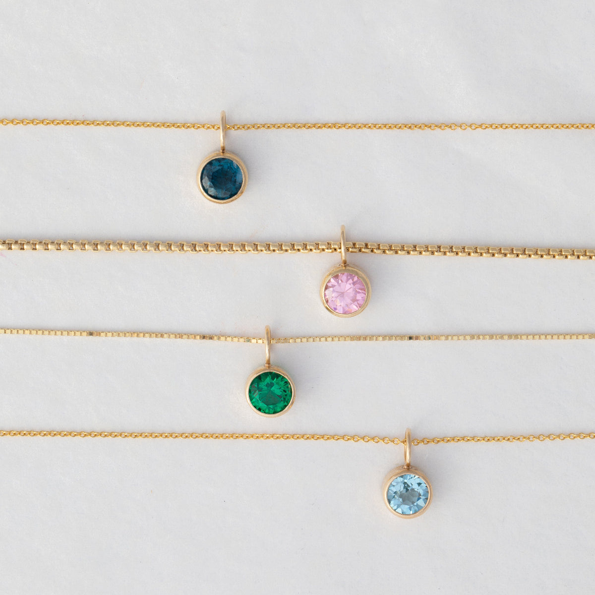 A Signature Cable Chain with a Color Pop Pendant in Blue (London Topaz), Box Chain with a Color Pop Pendant in Light Pink (Pink Tourmaline), The Slide Chain with a Color Pop Pendant in Green (Lab Grown Emerald) and a Signature Cable Chain with a Color Pop Pendant in Light Blue (Aquamarine) against a light grey background. 