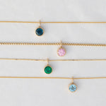 A Signature Cable Chain with a Color Pop Pendant in Blue (London Topaz), Box Chain with a Color Pop Pendant in Light Pink (Pink Tourmaline), The Slide Chain with a Color Pop Pendant in Green (Lab Grown Emerald) and a Signature Cable Chain with a Color Pop Pendant in Light Blue (Aquamarine) against a light grey background. 
