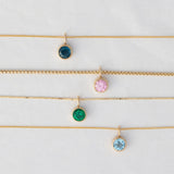 A Signature Cable Chain with a Color Pop Pendant in Blue (London Topaz), Box Chain with a Color Pop Pendant in Light Pink (Pink Tourmaline), The Slide Chain with a Color Pop Pendant in Green (Lab Grown Emerald) and a Signature Cable Chain with a Color Pop Pendant in Light Blue (Aquamarine) against a light grey background. 