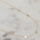 A Custom Gold Necklace in yellow gold with seven charms, the letters A and T, a heart, two birthstones, an om and a pavé letter N, on a marble surface.