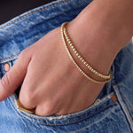 A hand in a front blue jean pocket wearing 
a Classic Bubble Bracelet and a Down the Line Bracelet, both in yellow gold. 