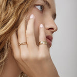 Close-up of a woman's hand wearing wearing a Perla Pinky Ring and a Criss-Cross Diamond Duo Ring in yellow gold, with her blonde hair visible against a light grey background. 