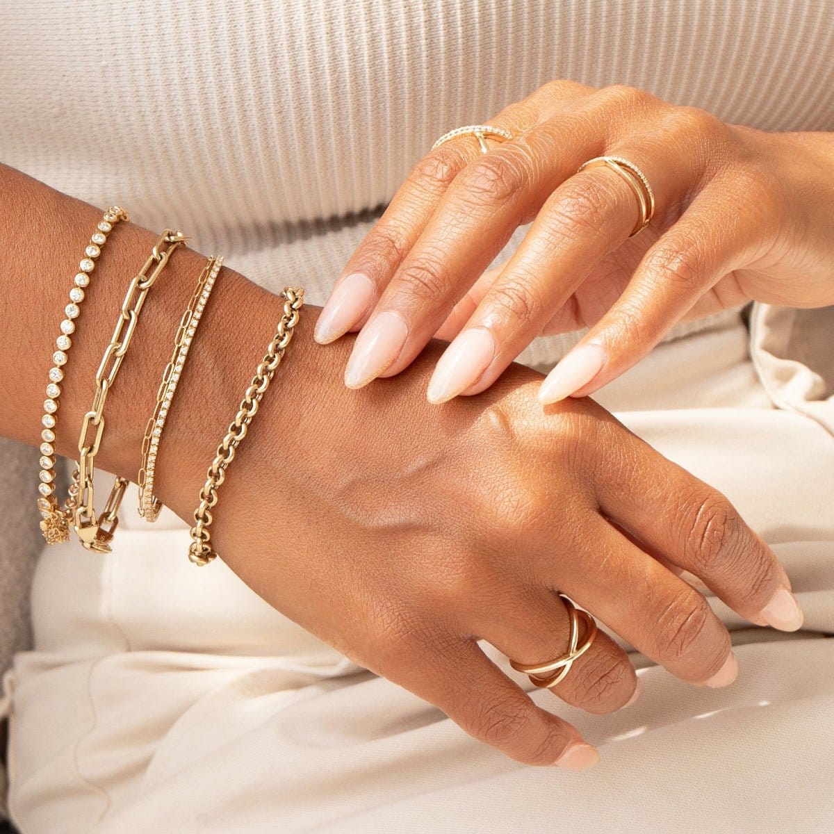 A close-up of a woman’s hands wearing a Love Set Match Bracelet, a Down the Line Bracelet, an Element Long Link Bracelet, an Element Short Link Bracelet and a Rolo Bracelet, as well as a Criss-Cross Duo Ring, a Criss-Cross Diamond Duo Ring and a Criss-Cross Diamond Daze Ring, all in yellow gold, styled in neutral clothing.