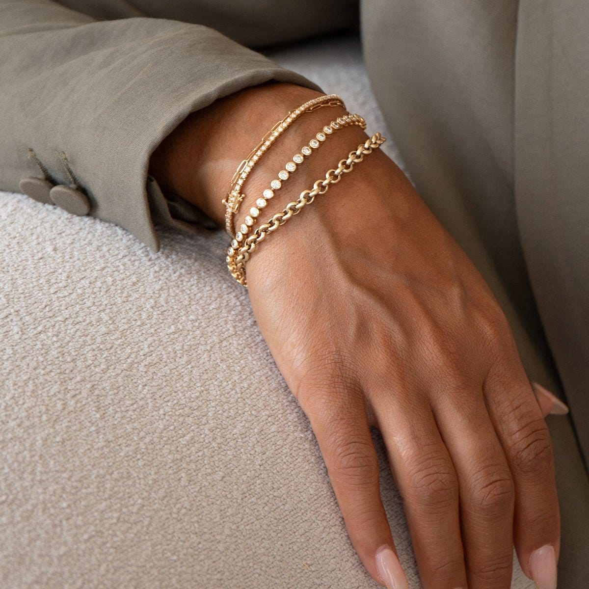 A close-up of a woman’s hand wearing an Element Short Link Bracelet, a Down the Line Bracelet, a Love Set Match Bracelet and a Rolo Bracelet, all in yellow gold, styled in a sage green jacket against a neutral background.