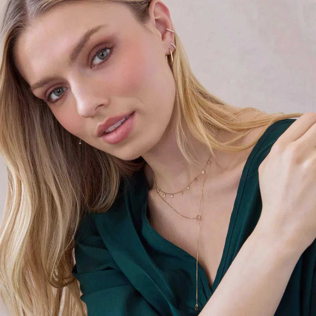 A blonde woman wearing a Stardust Diamond Necklace, a Gold Lariat Necklace, an Oval Hoop, a Pavé Double Ear Cuff and a Pavé Ear Cuff, styled in an emerald green top, against an off-white background. 