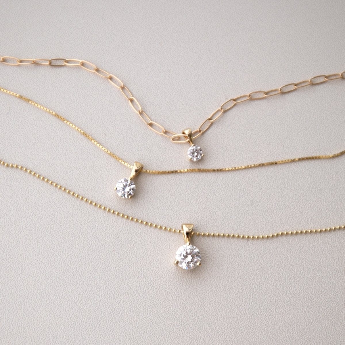 A Duchess Diamond Pendant on an Element Long Link Chain, a Princess Diamond Pendant on The Slide Chain and a Queen Diamond Pendant on a bead Chain in yellow gold against a light grey background. 