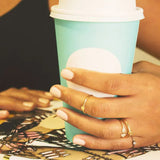 A woman's hand wearing several rings, including a Geo Stacking Ring | Triangle and Petal Ring, with a black tank top holding a coffee cup. 