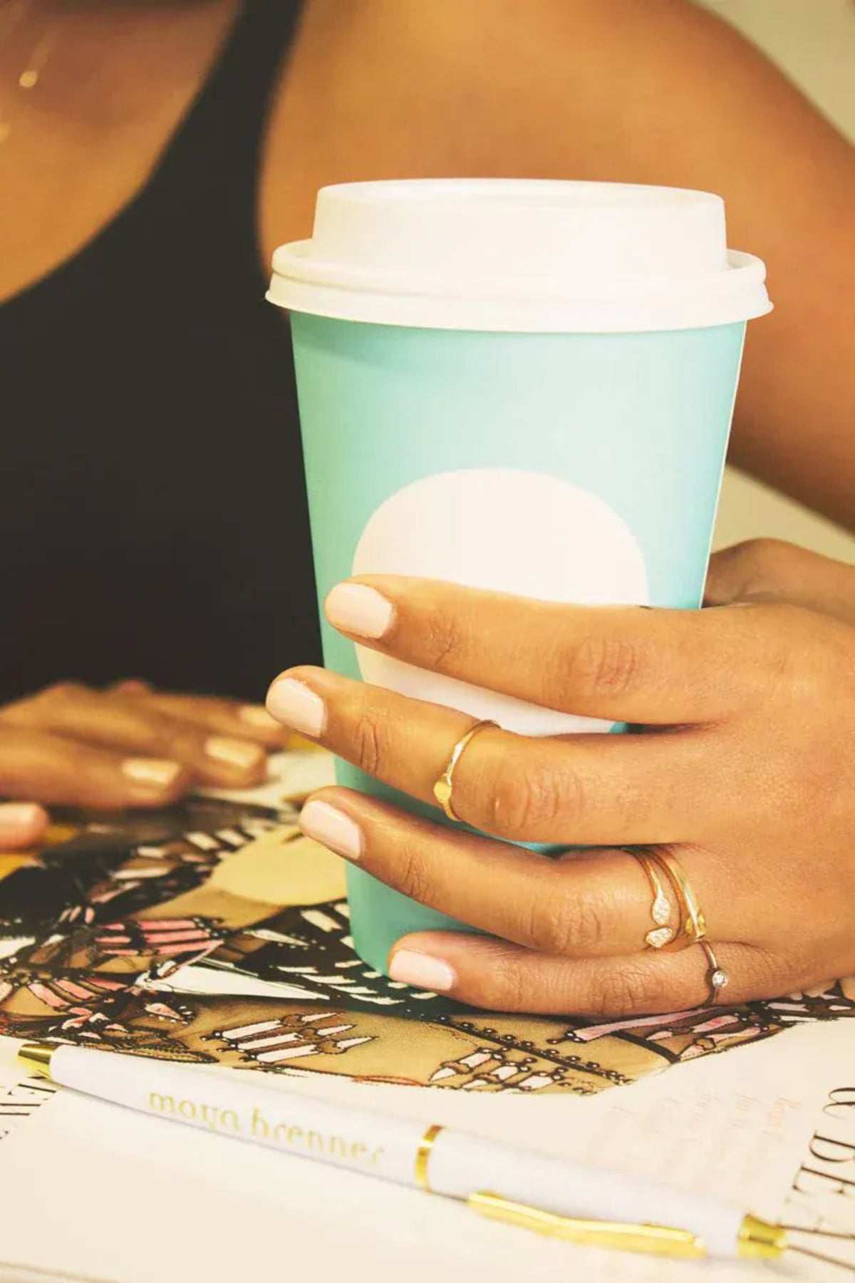 A woman's hand wearing several rings, including a Geo Stacking Ring | Triangle and Petal Ring, with a black tank top holding a coffee cup. 