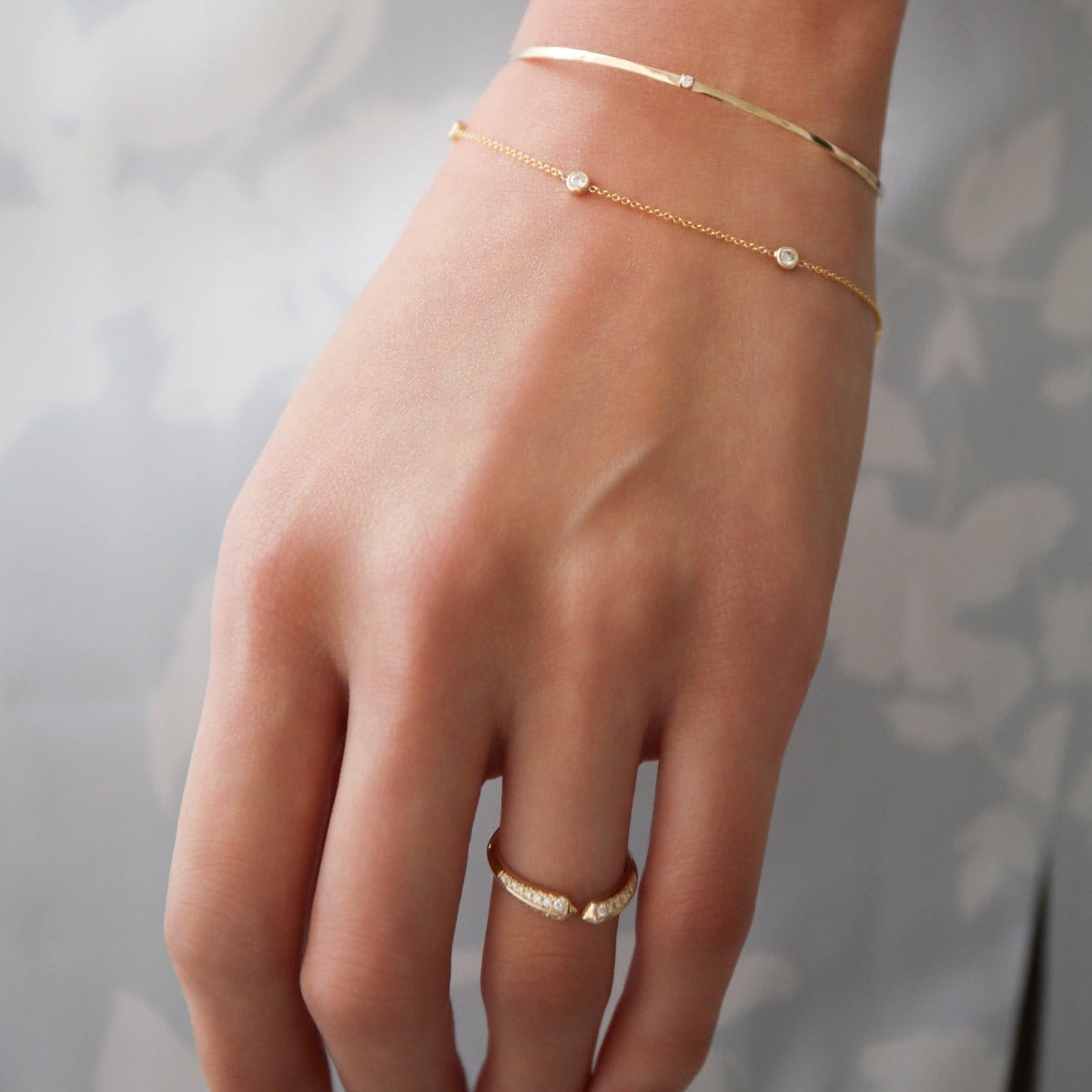 A close-up of a woman's hand wearing an Off the Cuff Bracelet, a Starstruck Diamond Bracelet and an Open Diamond Ring, against a white and light grey floral background.