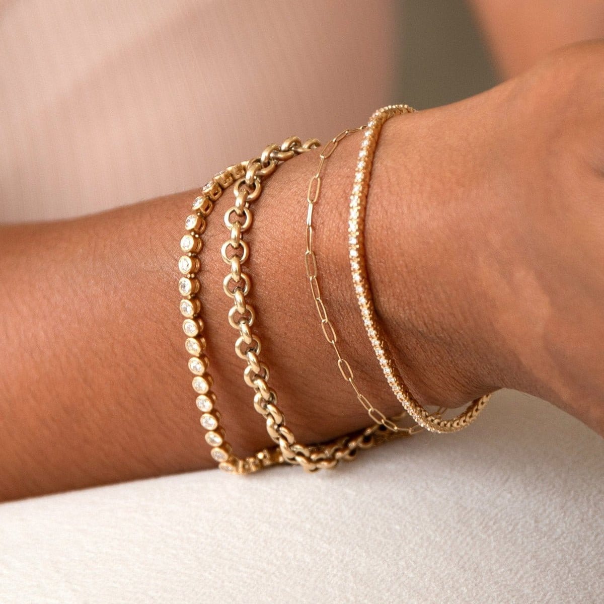 A close up of a wrist wearing a Love Set Match Bracelet, a Rolo Bracelet, an Element Short Link Bracelet and a Down the Line Bracelet, all in yellow gold against a neutral background. 
