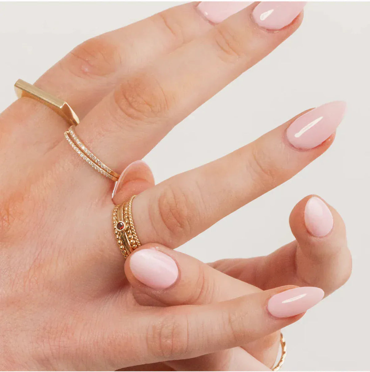 Hand wearing Delicate Trio Stacking Rings, a Birthstone Ring, two Pavé Diamond Rings,  Geo Stacking Ring Square, with pink nail polish on a light grey background.