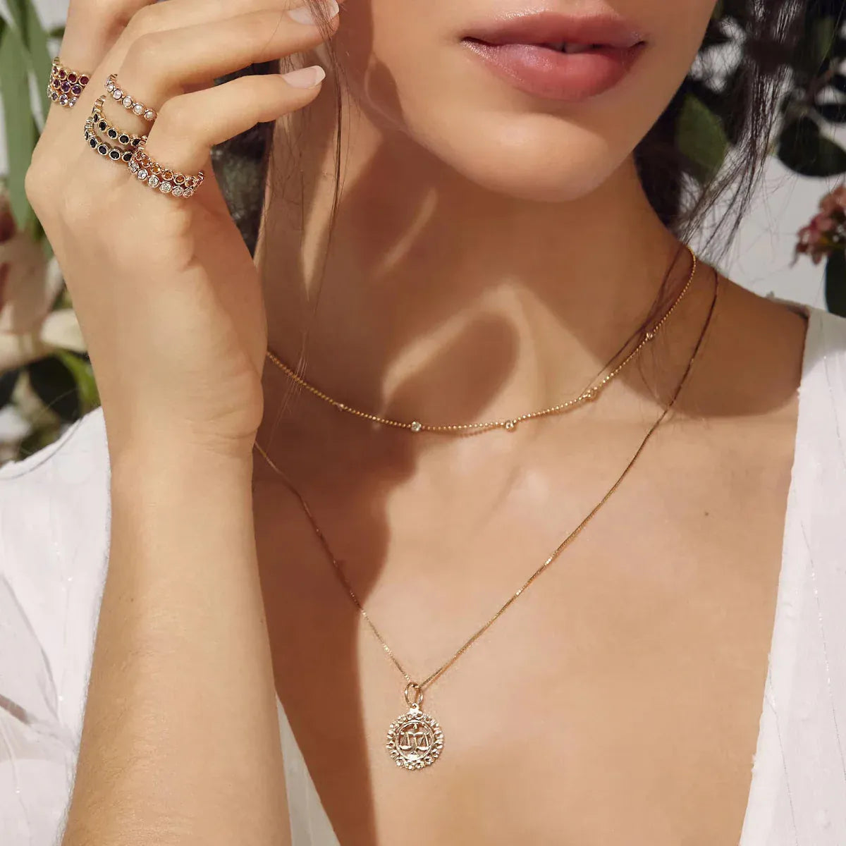 Close-up of a woman's décolletage wearing a yellow gold Quintet Birthstone Layering Necklace and a Zodiac Necklace  - Libra, her hand wearing several Bouquet Rings, with a white top and a blurred leafy background.