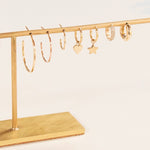 Sleek & Chic Hoops in Large, Medium and Small, Charmed I'm Sure Hoops in Star and Heart, a Pave Huggie and a Gold Huggie, all in yellow gold, displayed on a gold jewellery stand against a light background. 