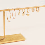 Sleek & Chic Hoops in Large, Medium and Small, Charmed I'm Sure Hoops in Star and Heart, a Pave Huggie and a Gold Huggie, all in yellow gold, displayed on a gold jewellery stand against a light background. 