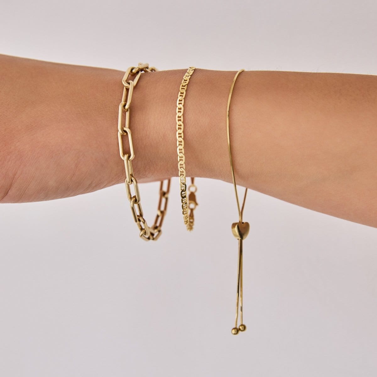 A close-up of an arm wearing The Slide Heart Bracelet, a Bold Link Bracelet and an Overboard Bracelet, all in yellow gold against a light grey background.