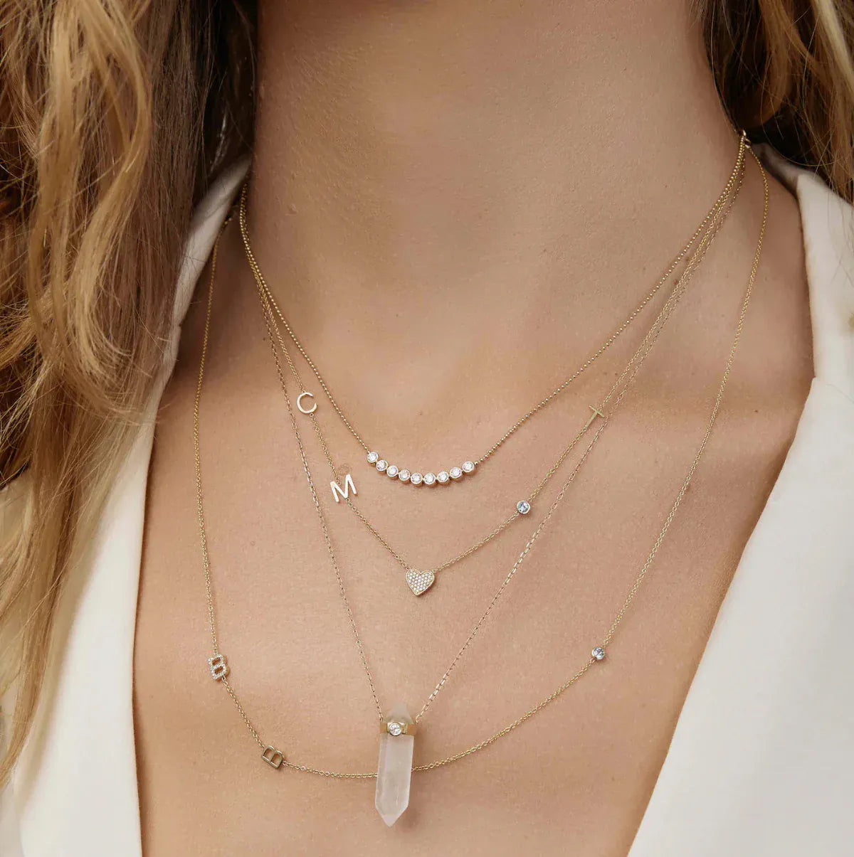 A close-up of a woman’s neck wearing four yellow gold necklaces - two Custom Gold Necklaces (one with three charms and the other with four charms), the Clarity Retreat Necklace and the Diamond Arc Layering Necklace, with a cream top.